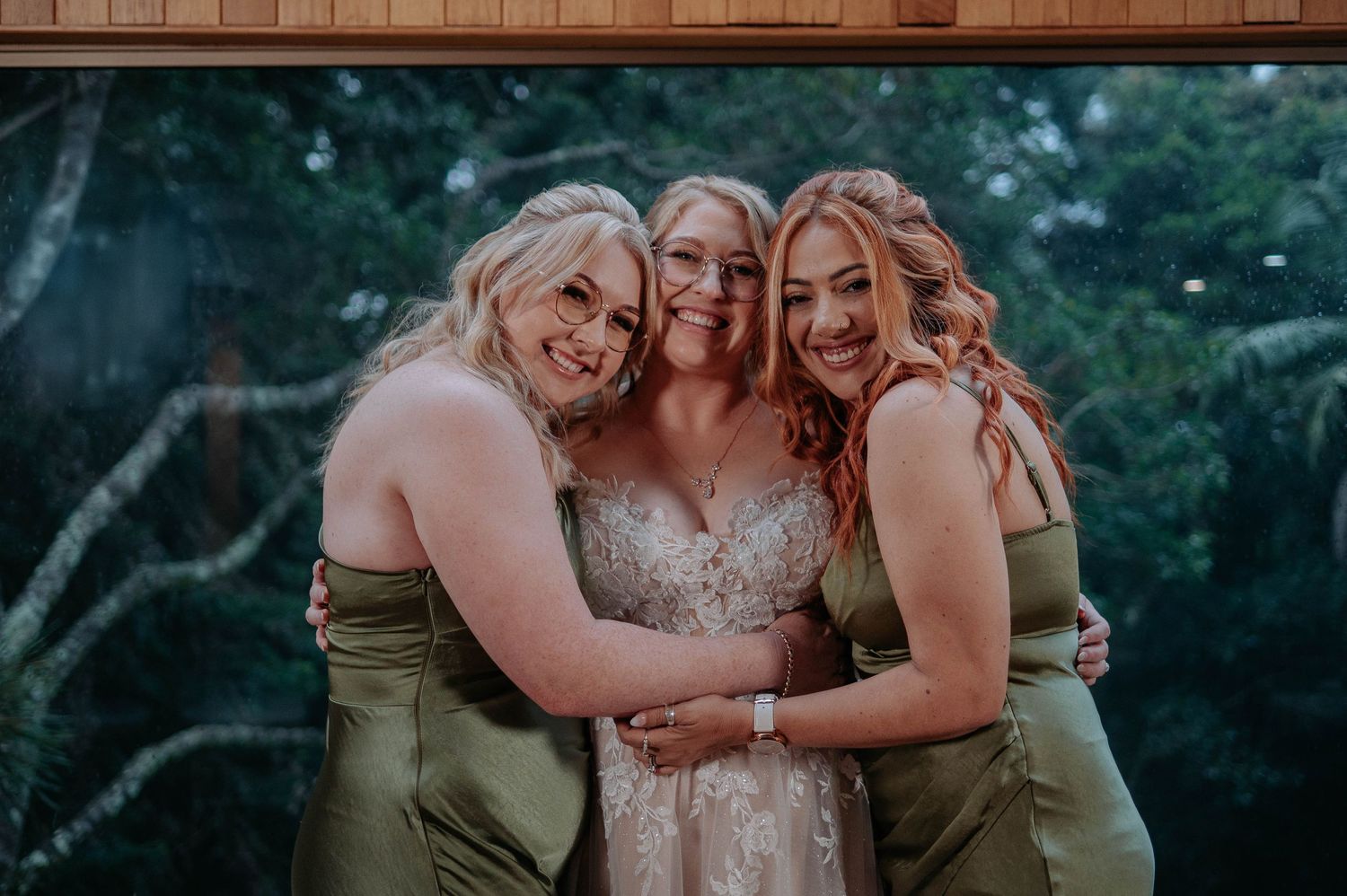 Three women in matching sage dresses share embrace during wedding celebration.