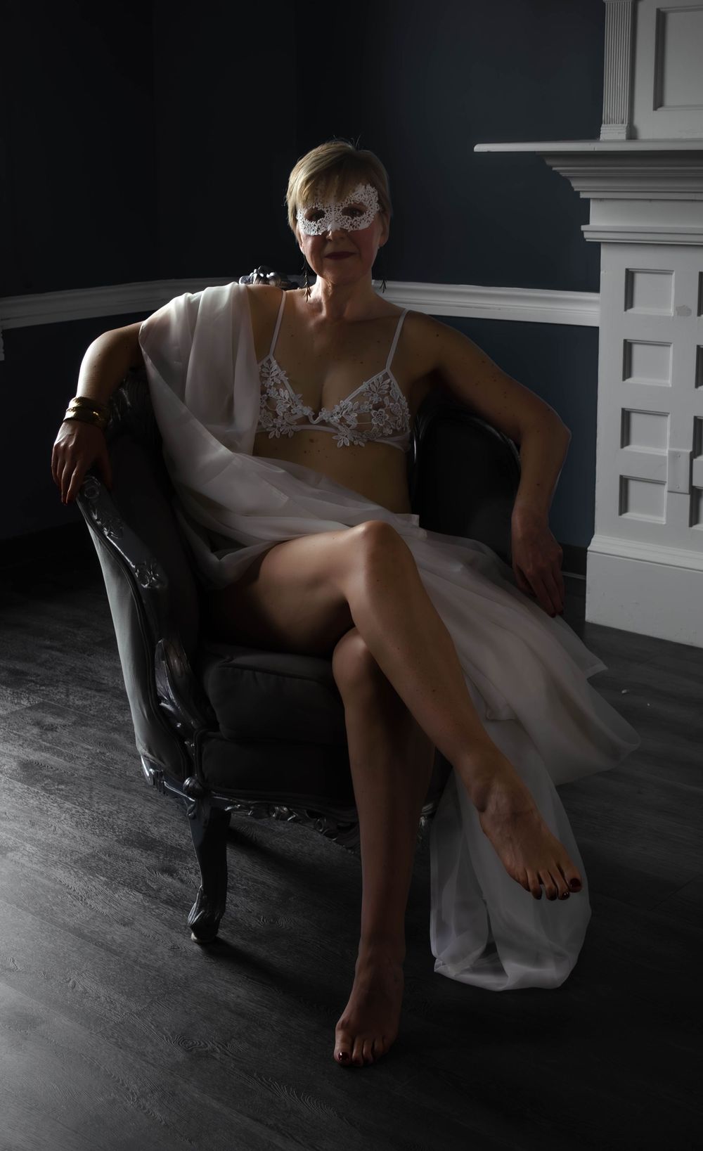 Figure in white lingerie and masquerade mask poses elegantly on dark vintage chair against moody lighting.