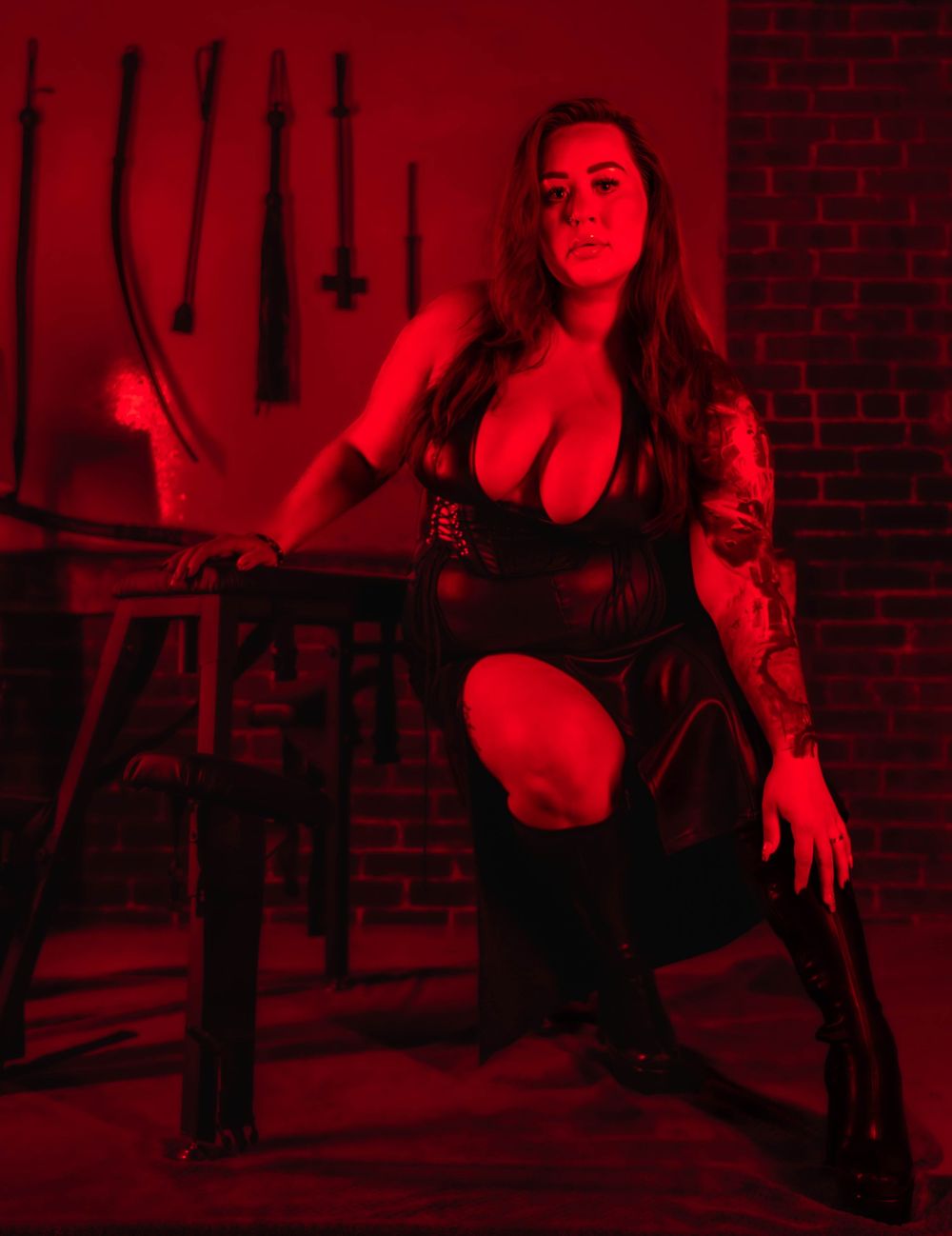 A person in black attire poses dramatically in a red-lit room with brick walls and hanging implements.