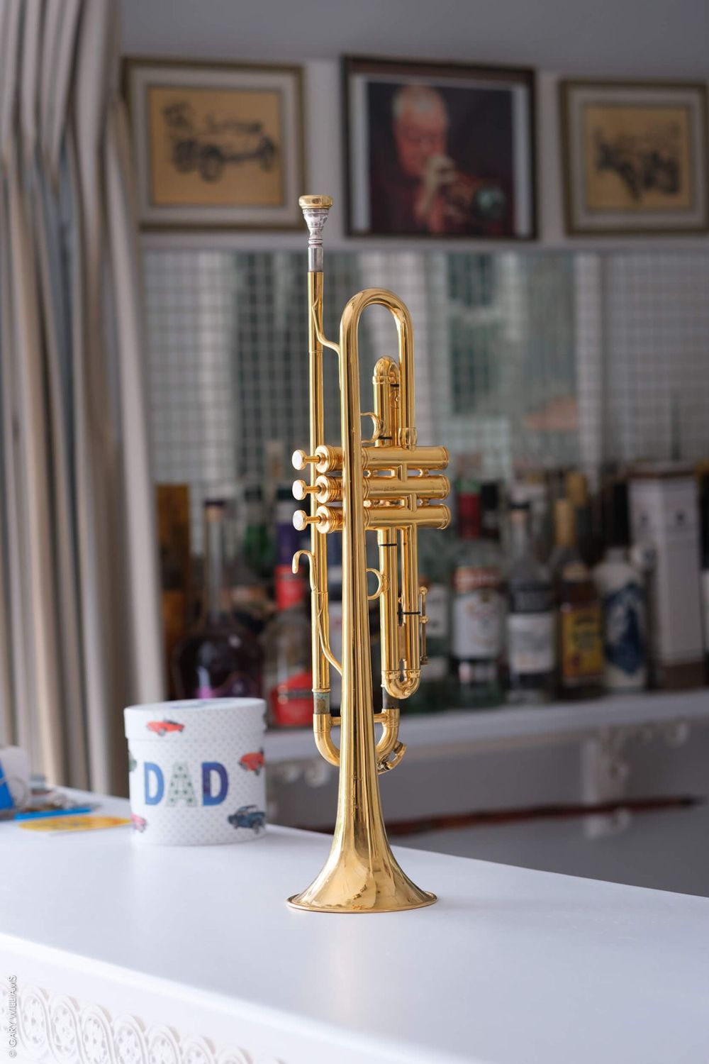 A shiny brass trumpet stands upright on a white surface with a blurred background of framed pictures and kitchen items.