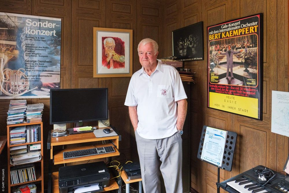 A white-haired person stands in a small wood-paneled office with posters and audio equipment visible on the walls.