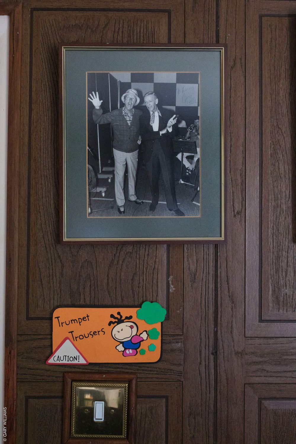 A framed vintage black and white photograph hangs on a wooden wall above a colorful trumpet player sign.