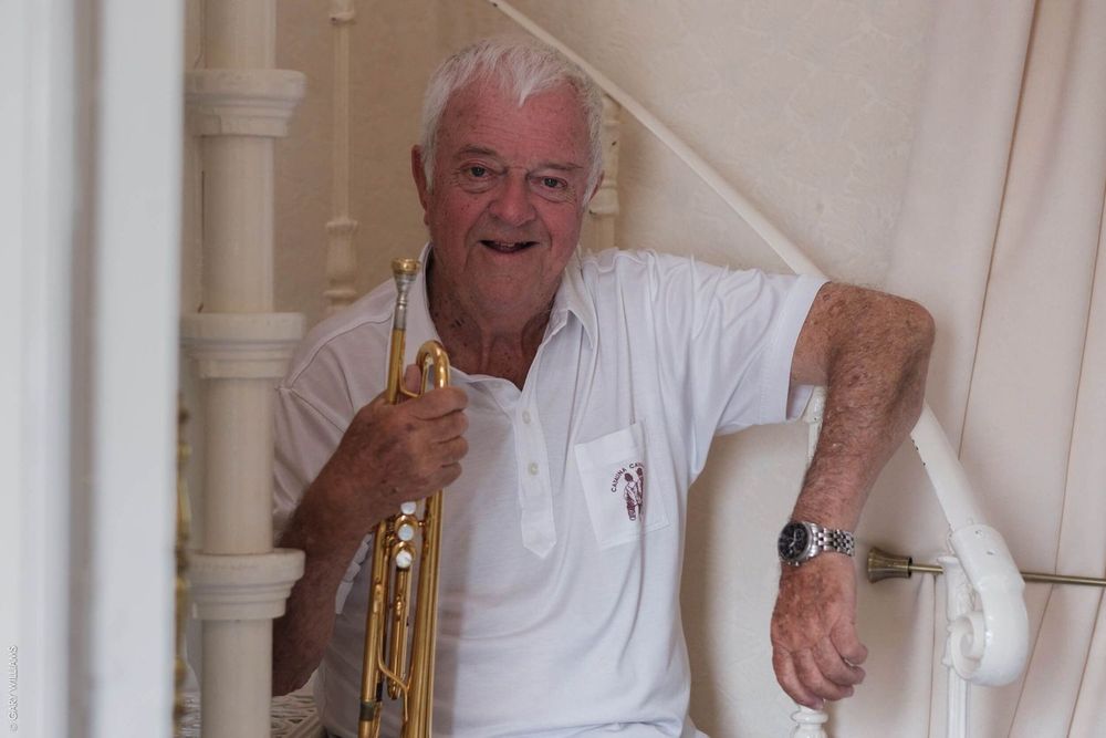 A musician in a white polo shirt holds a brass trumpet while leaning against a white pillar indoors.