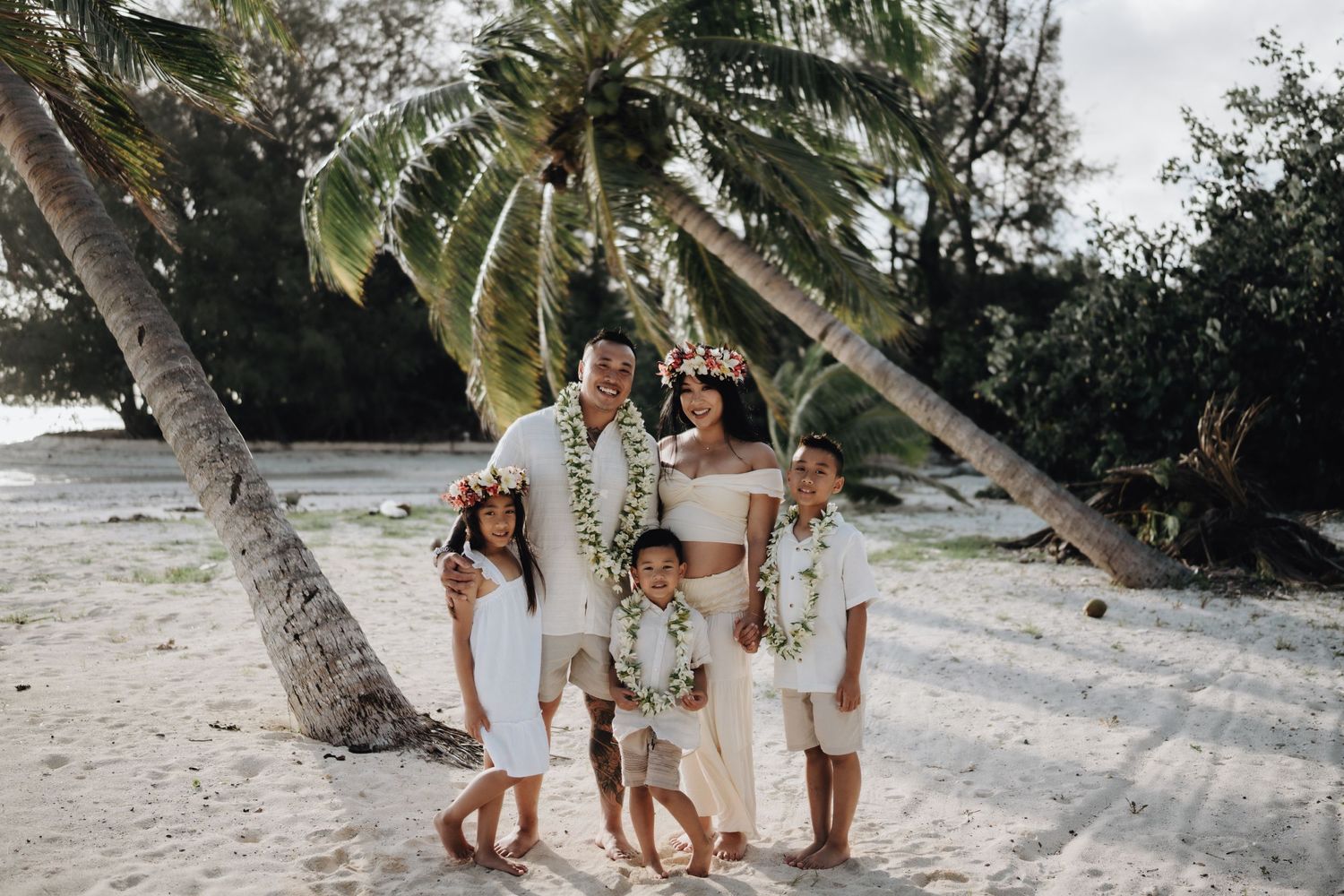 A family wearing white outfits poses together on a tropical beach with palm trees and ocean waves in the background.