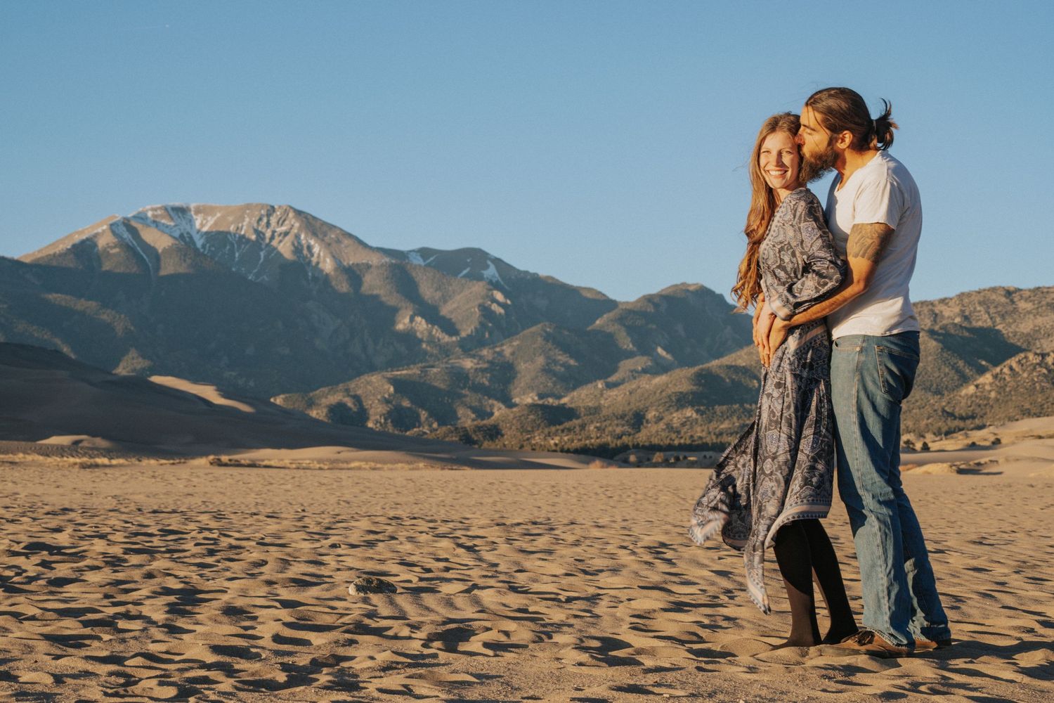 A couple embraces while standing on sand dunes with majestic mountains in the background during golden hour.