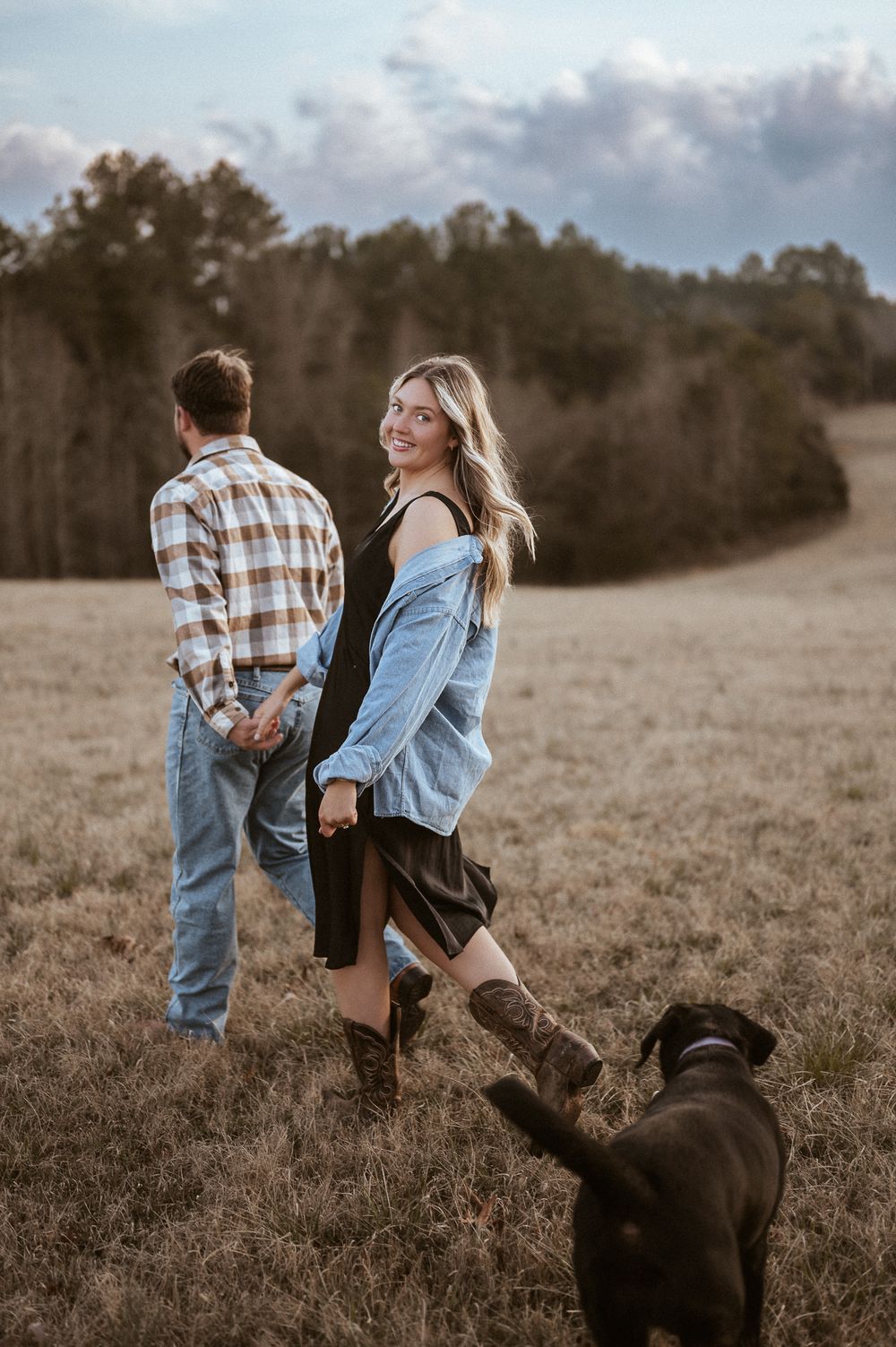 Couple posing in field at sunset with dog watching from below.