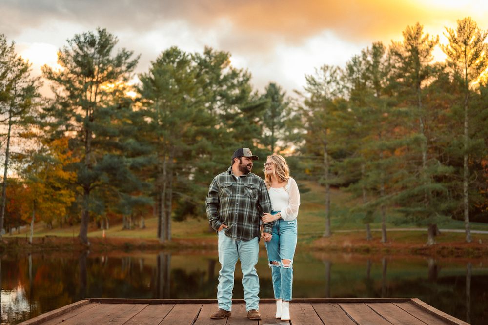 Couple stands together on wooden dock overlooking autumn lake with pine trees at sunset.