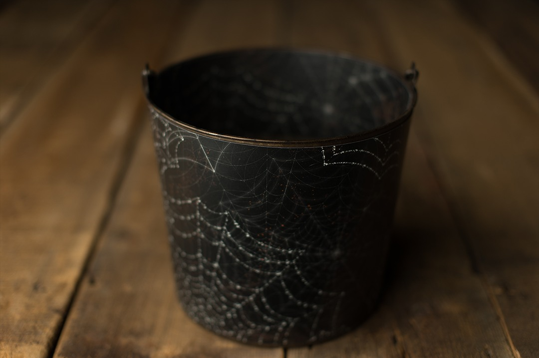 A dark black metal bucket with etched spiderweb design sitting on a rustic wooden surface.