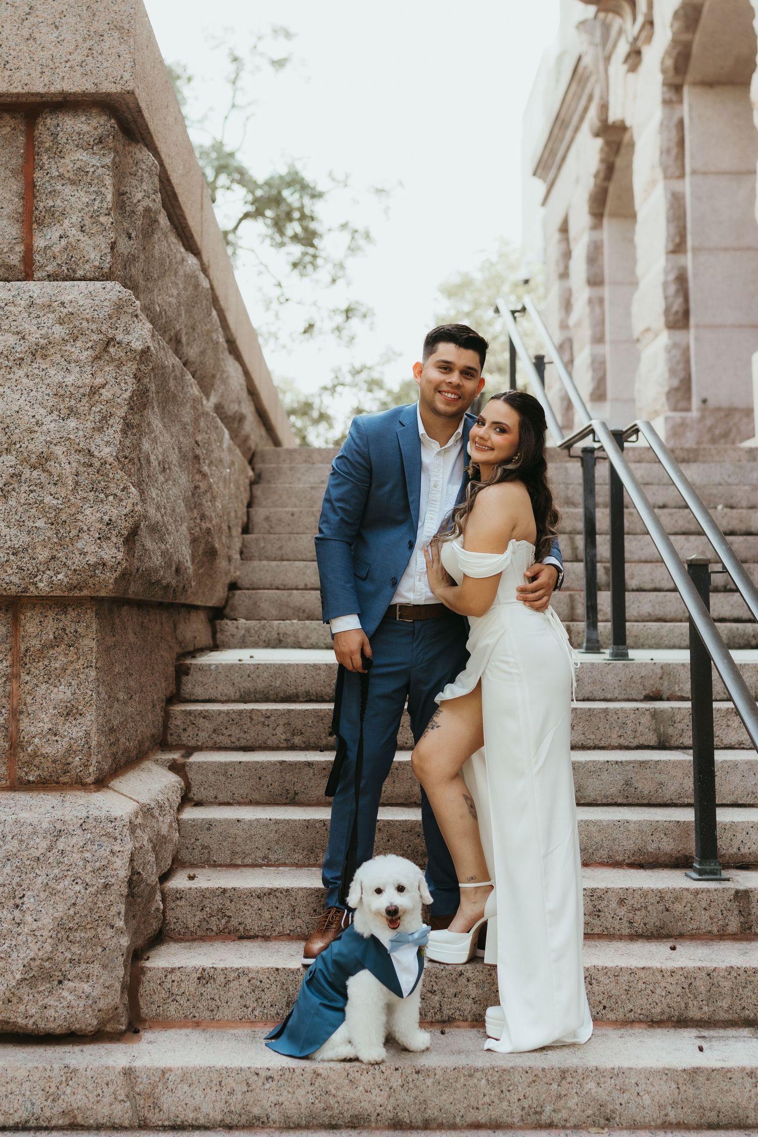 A couple sits on stone steps with their white dog during an engagement photo session.