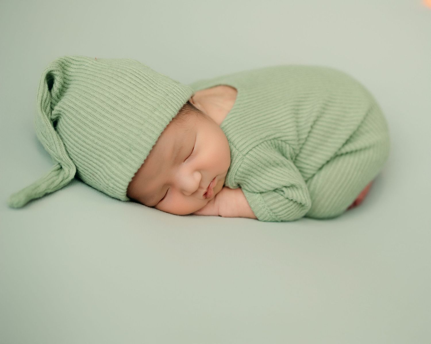 A sleeping newborn curled up in soft mint green knit clothing and matching beanie on light green backdrop.
