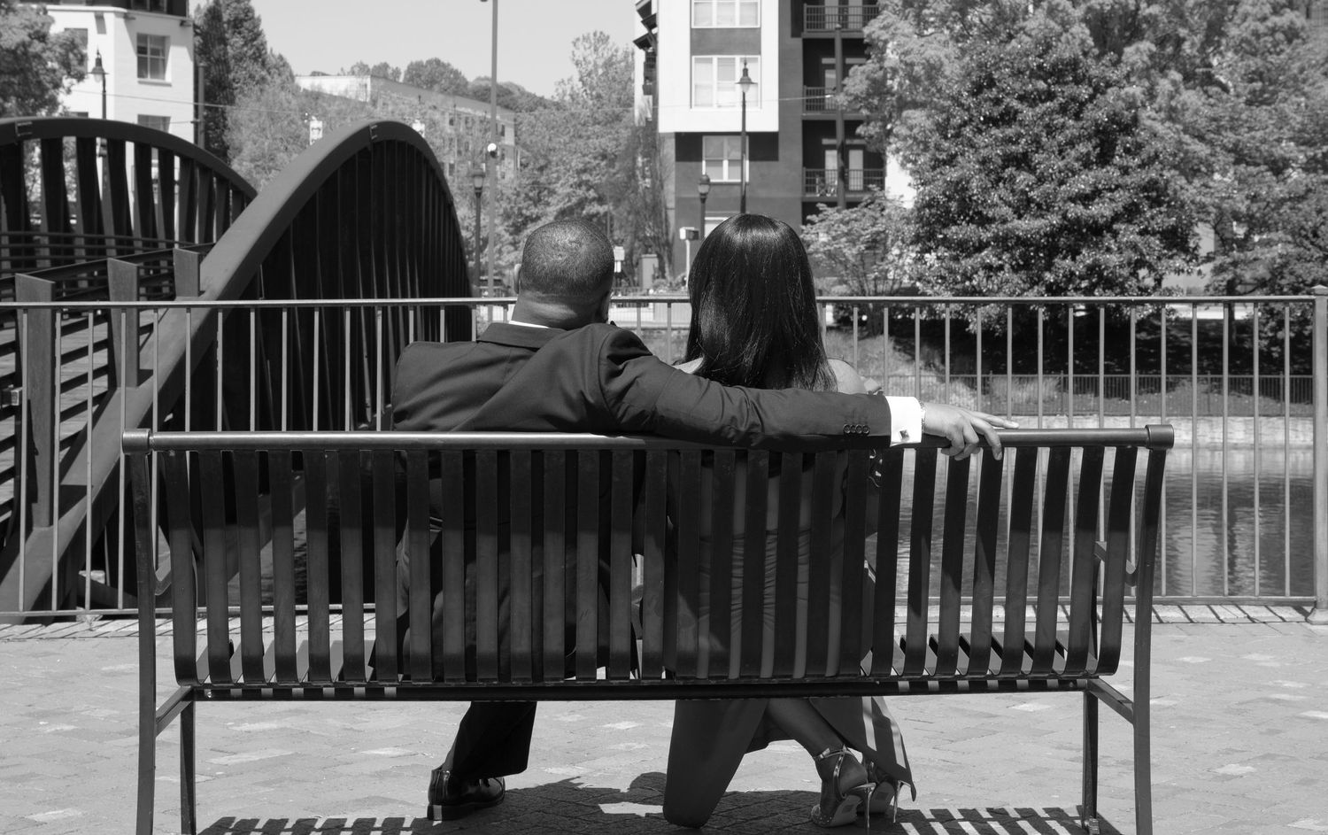Two people sit together on a wooden bench overlooking a canal with an arched bridge in black and white.