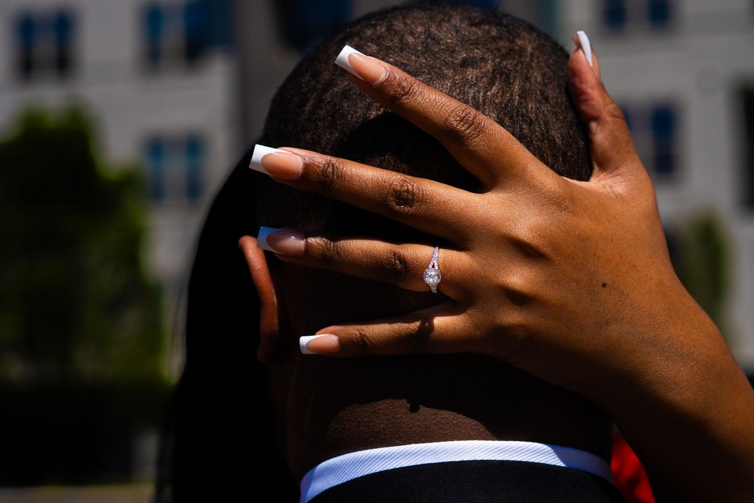 Hand with an engagement ring reaches toward someone's head against a blurred building background in sunlight.