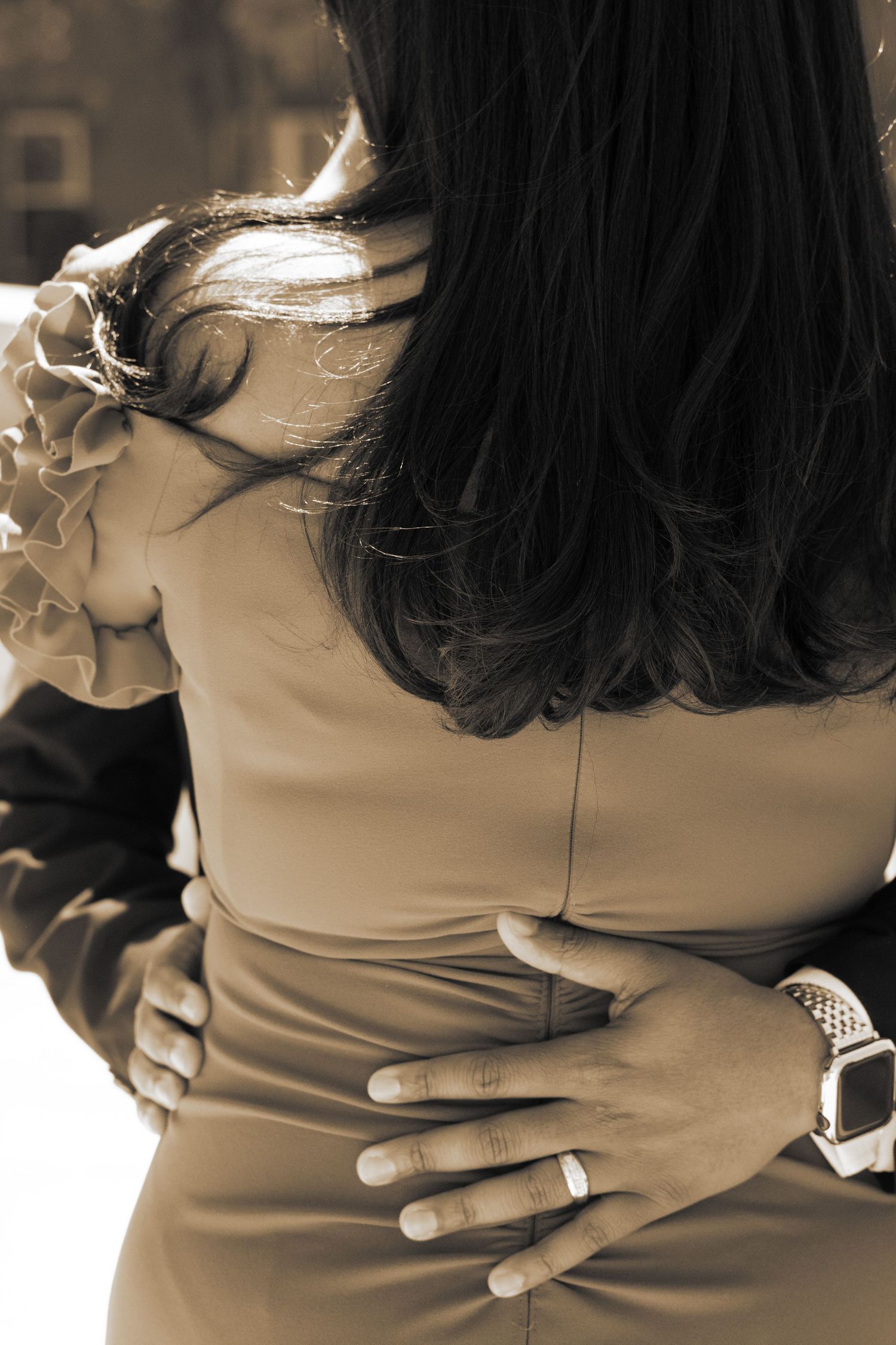 A close-up sepia toned photo showing hands embracing around a fitted dress waist with elegant jewelry details.