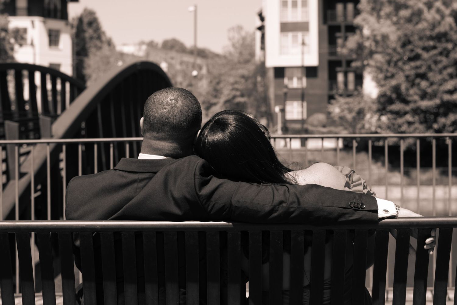 A couple sits close together on a park bench in a black and white photograph overlooking a canal.
