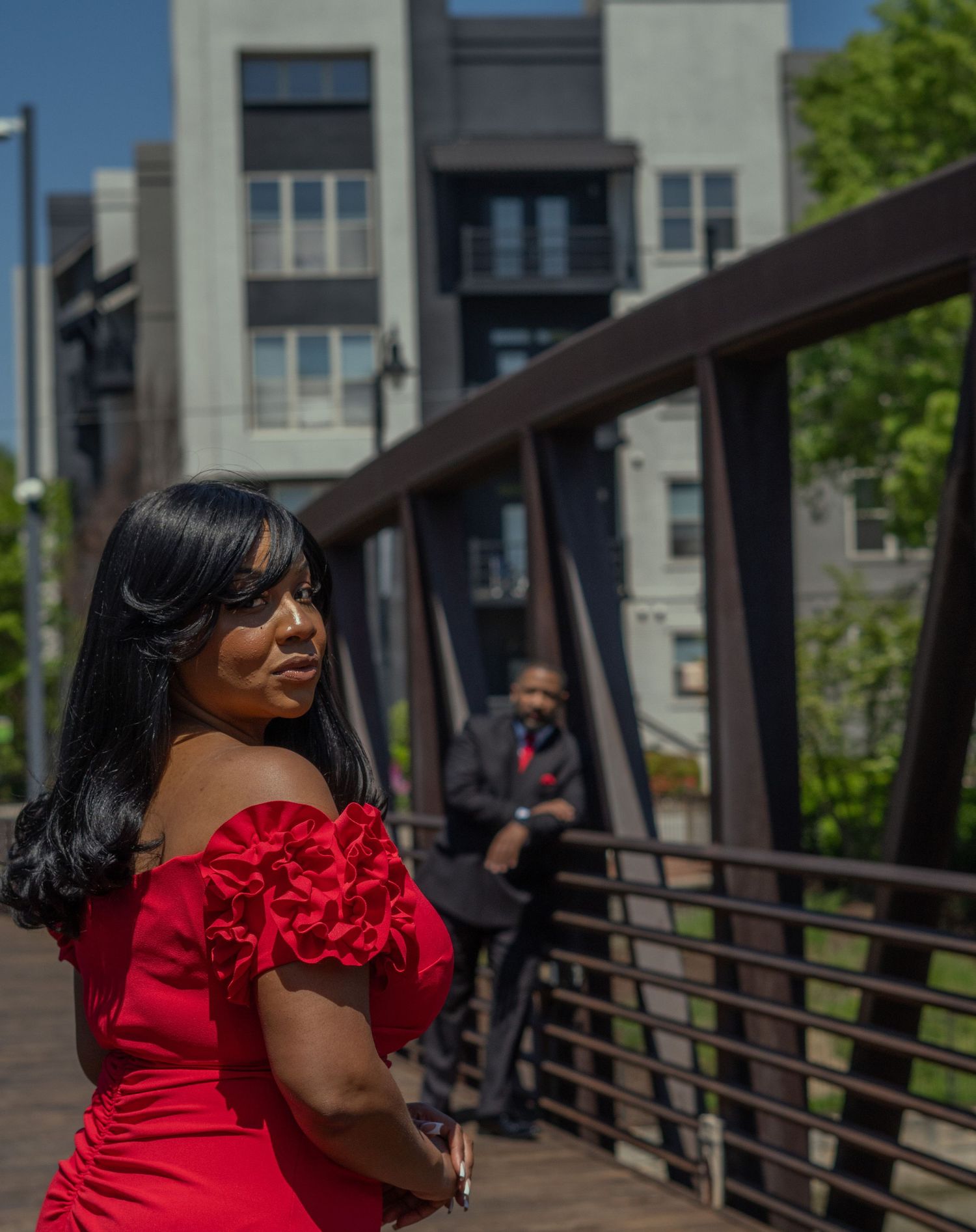 Person in a vibrant red ruffled dress stands on a metal bridge with urban buildings in the background.