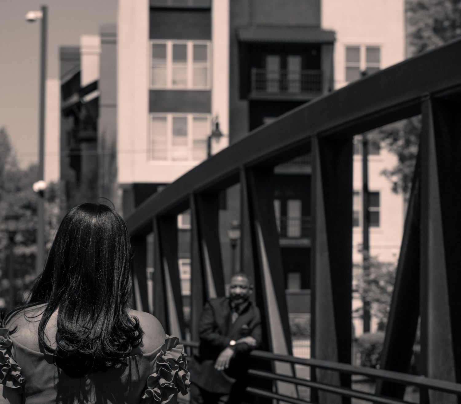 A black and white photo showing a person walking across a metal bridge with apartment buildings in the background.