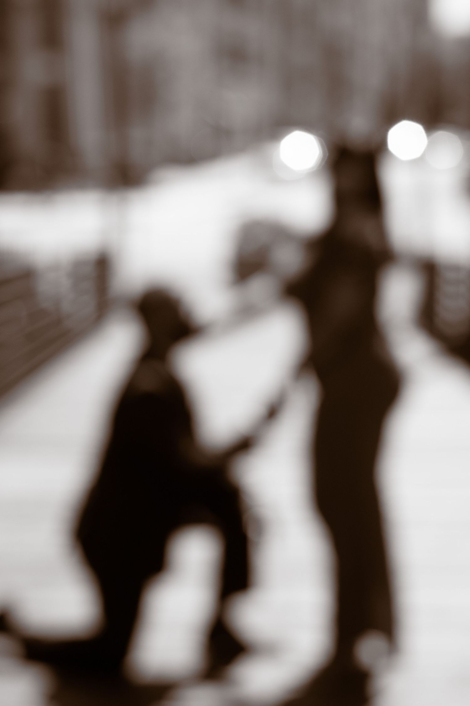 Blurry black and white photograph showing silhouettes of people walking on a city street with bokeh lights in background.