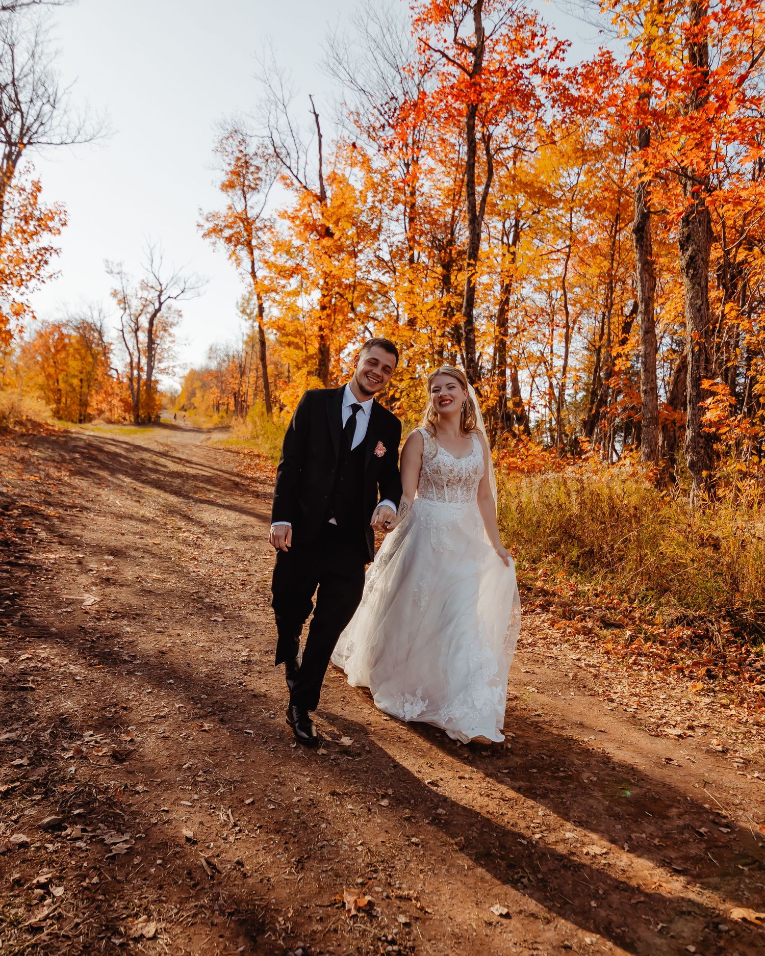 A couple walks hand in hand down a dirt path surrounded by vibrant orange fall foliage on their wedding day.