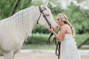 A beautiful white horse with a braided mane stands with someone in a bridal gown in an outdoor garden setting.