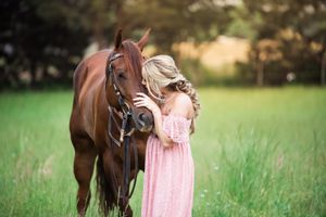A figure in a flowing pink dress stands with a chestnut brown horse in a sunlit green meadow.