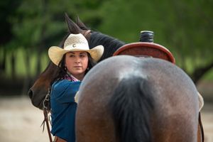 A horse rider wearing a cowboy hat and blue shirt peeks around the side of their brown horse during training.