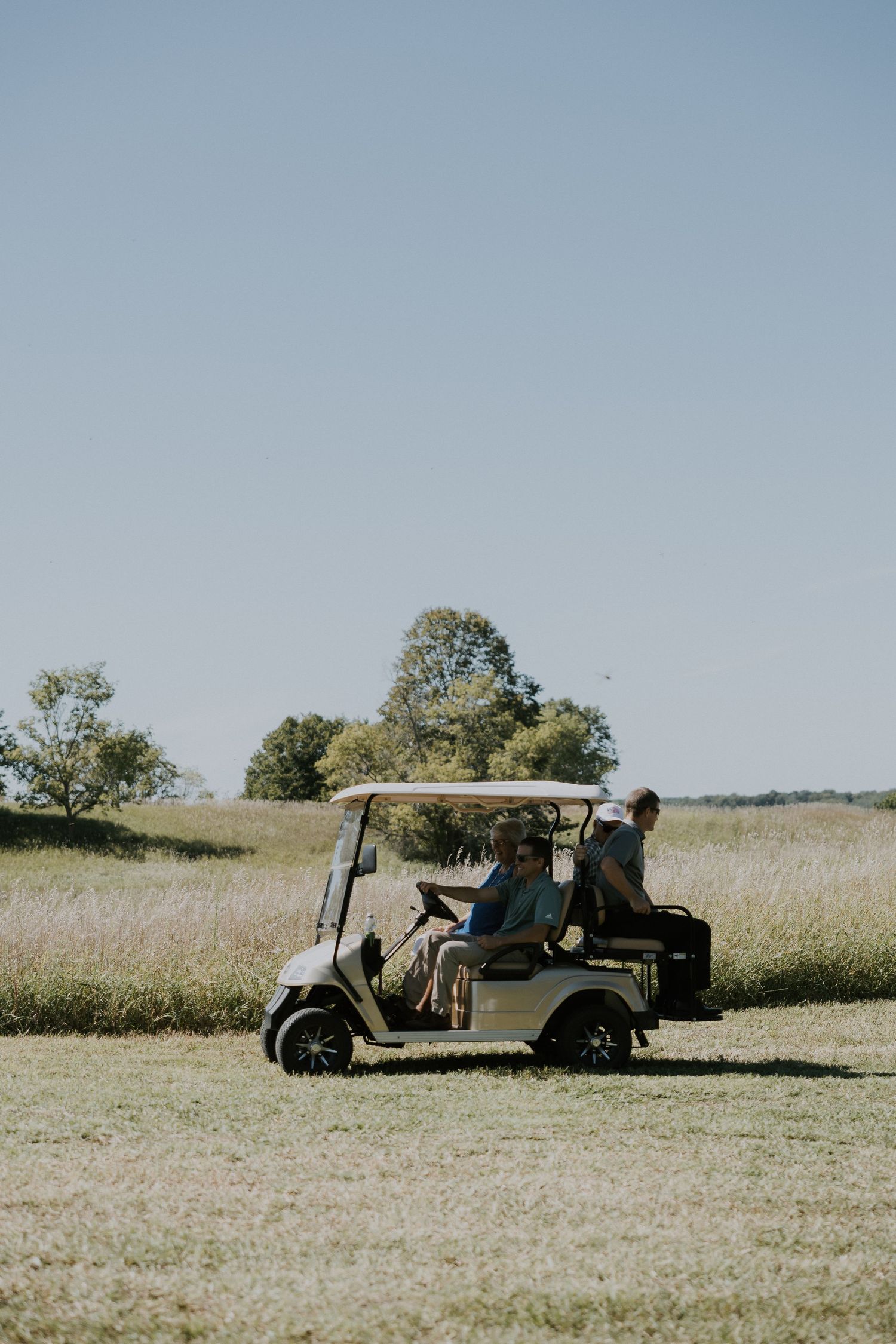 A golf cart drives across an open field with tall grass and trees in the background on a sunny day.