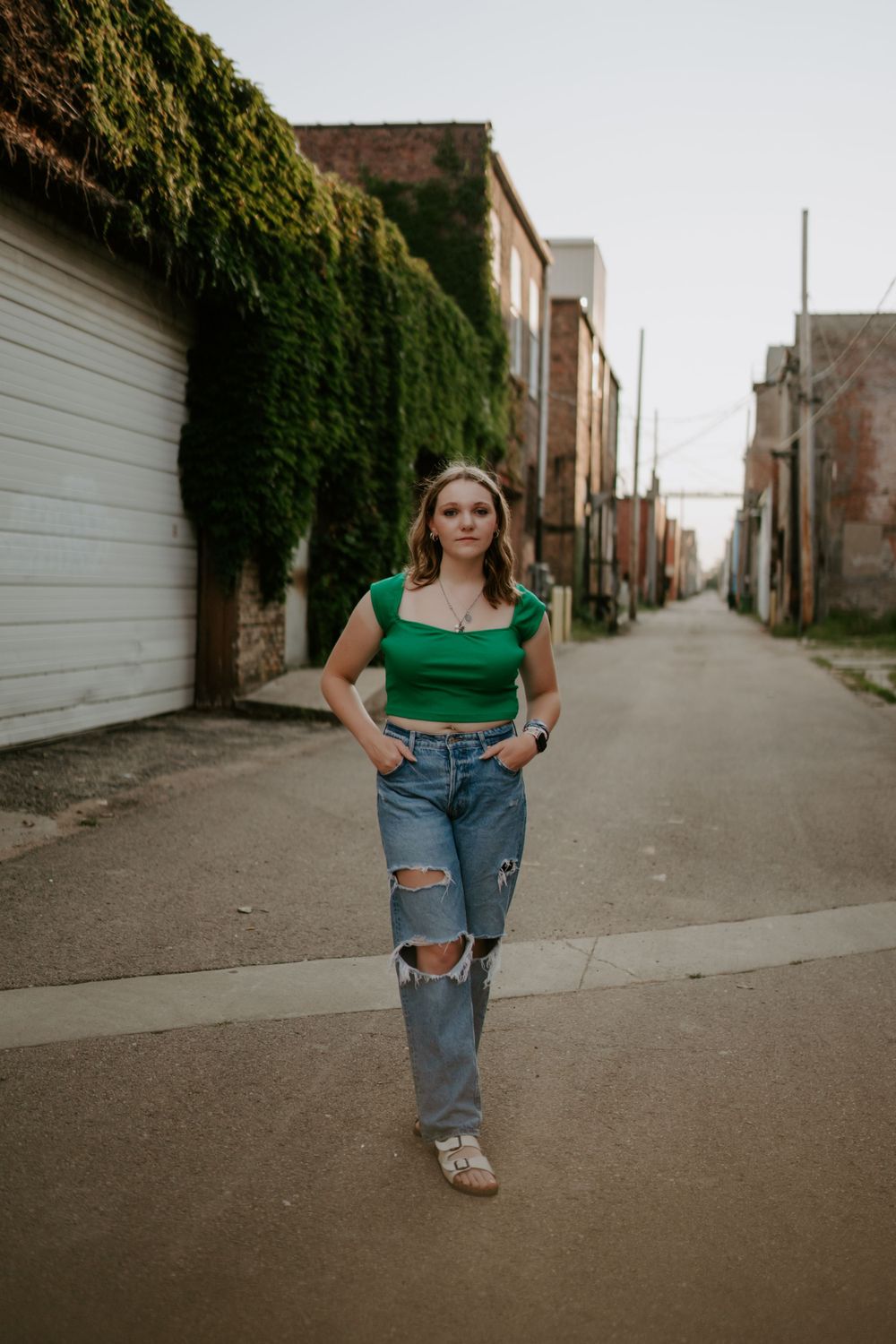 Urban alley photoshoot with ivy-covered walls featuring casual denim and green top outfit.