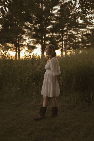 A person in a white dress stands in a field at sunset with trees silhouetted in the background.