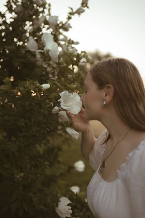 Someone in a white dress smells white flowers on a bush during golden hour.