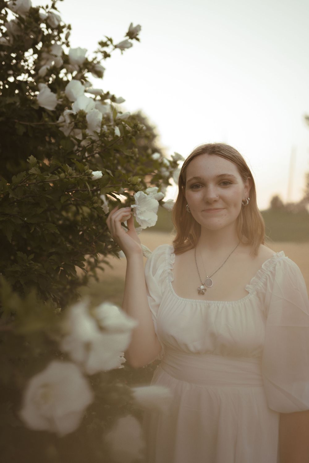 A person wearing a white dress poses near blooming flowers during sunset.