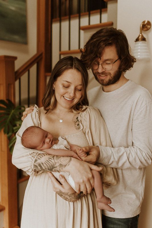 A young couple in casual attire stands indoors holding their sleeping newborn baby in a warm, intimate moment.