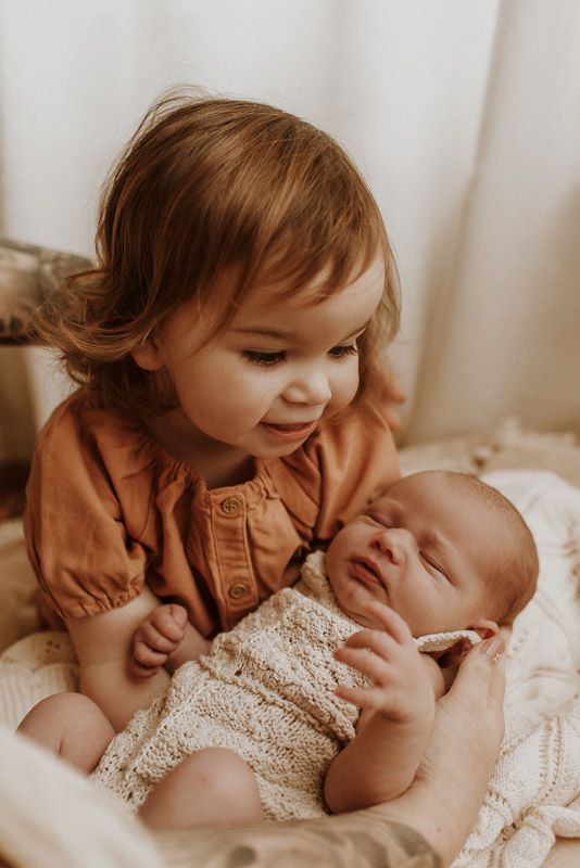 A young child in a brown shirt gazes lovingly at a sleeping newborn baby wearing white lace.