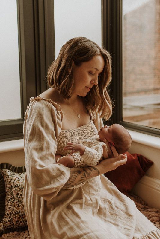 Mother in white dress holds and gazes lovingly at newborn baby while sitting by window in warm natural light.