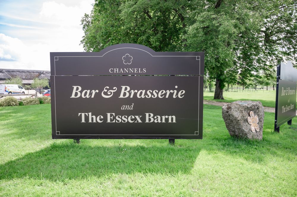 A black sign for Bar & Brasserie and The Essex Barn stands on a grassy lawn with trees in the background.