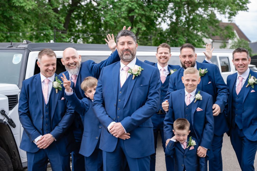 A wedding party in matching navy blue suits gather around a car for a fun group photo.