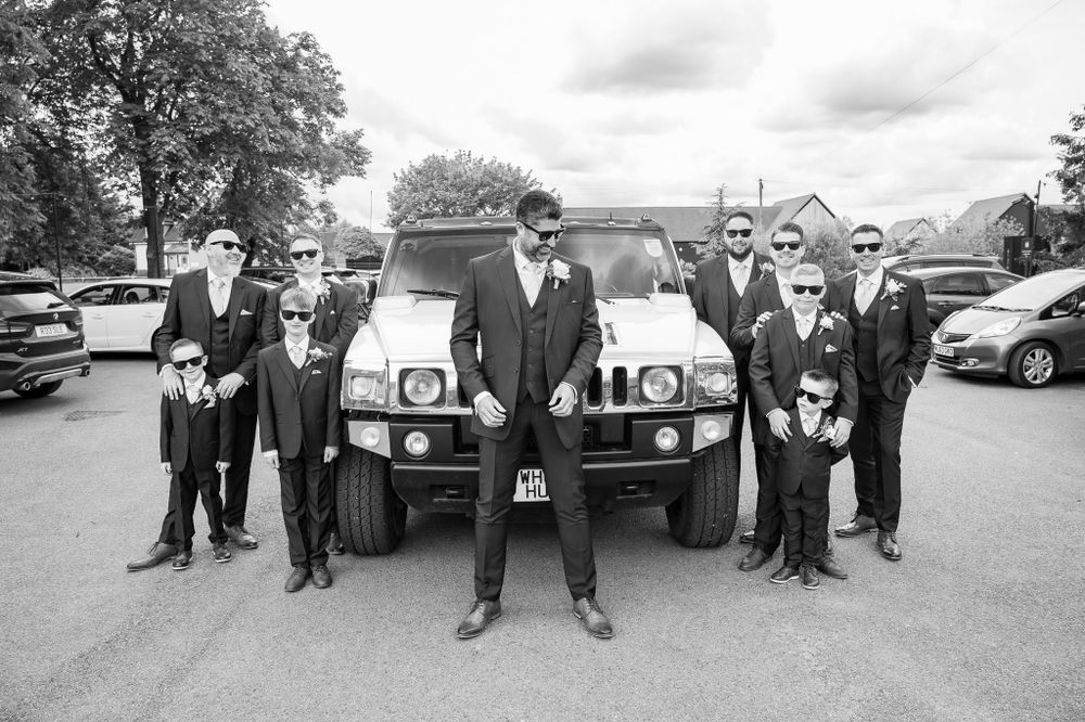 A wedding party poses in formal attire in front of a classic vehicle on a cloudy day.