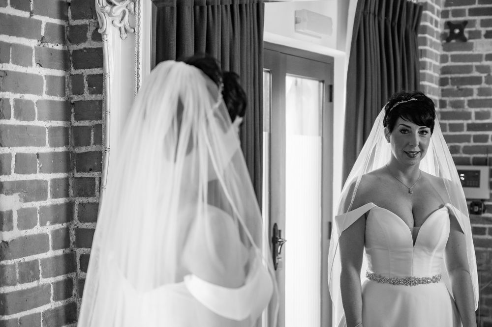 A bride in a white gown with a veil looks at her reflection in a mirror against exposed brick walls.