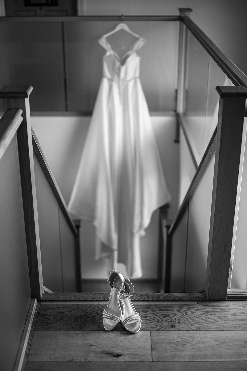 A white wedding dress hangs elegantly on a dark staircase with bridal shoes placed below in black and white photography.