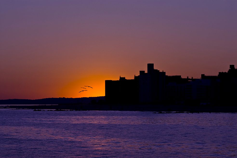 Dramatic sunset silhouettes a castle fortress along a purple-hued coastline with the sun dipping below the horizon.