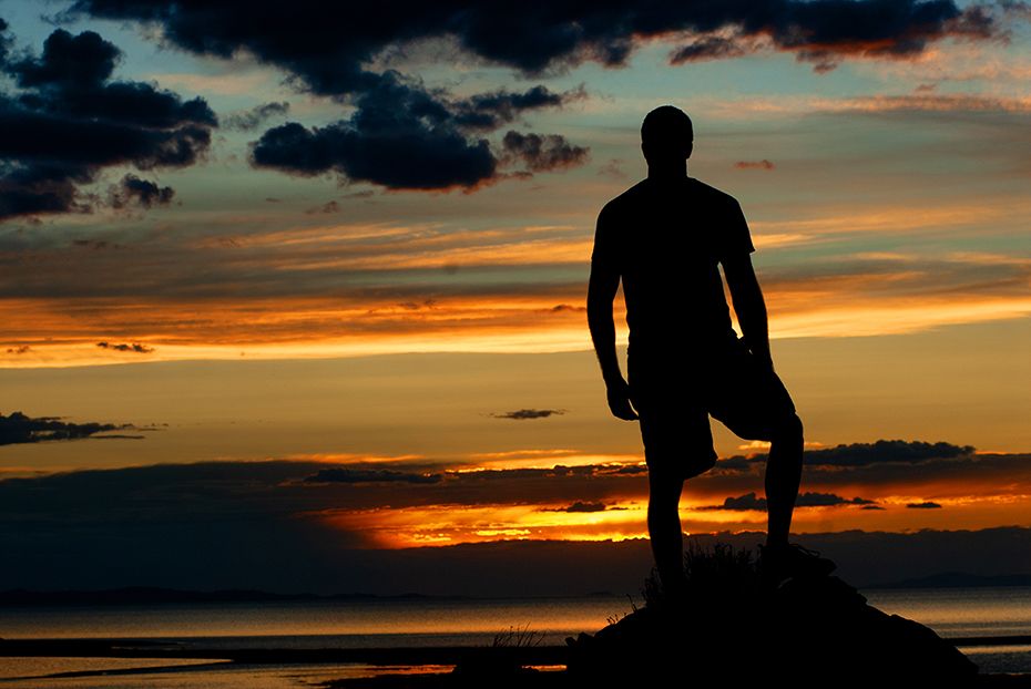 Silhouette against vibrant orange sunset sky stands on hill overlooking tranquil water.