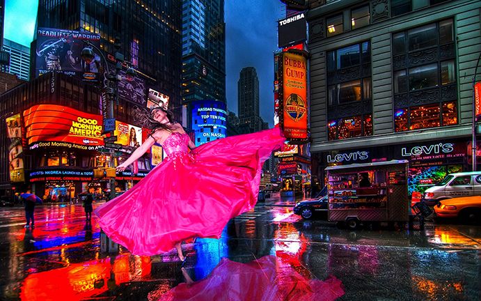 A woman in a vibrant pink dress reflects in wet streets of Times Square at night amidst neon lights and city buildings.