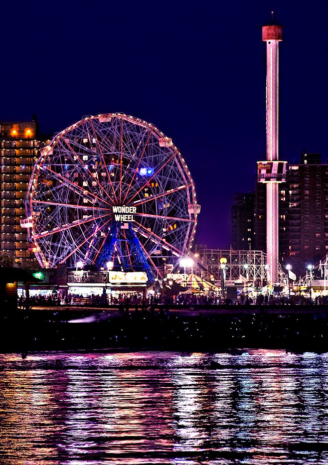 Illuminated Wonder Wheel and amusement rides at Coney Island reflect colorfully in the water at night.