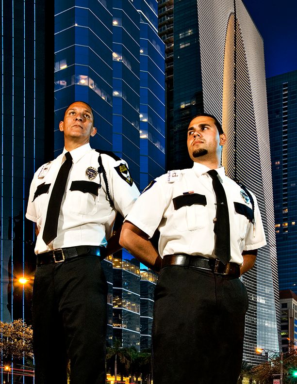 Two security guards in white uniforms stand at attention in front of modern glass skyscrapers at night.
