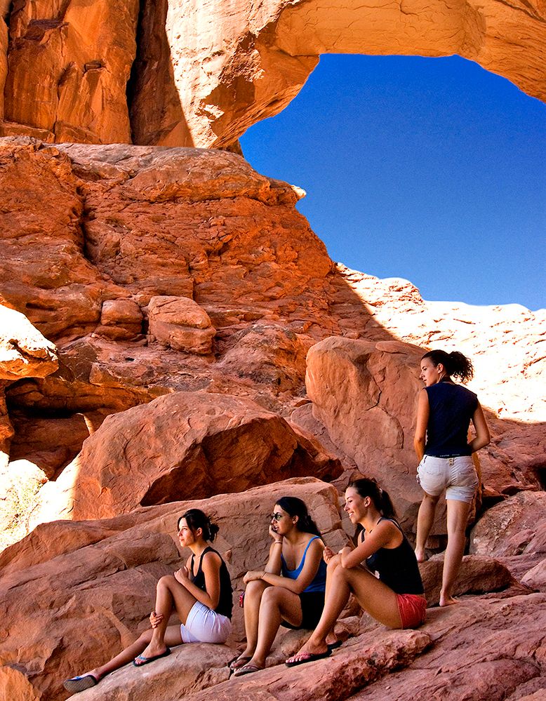 Hikers resting beneath a natural red rock arch formation in the desert with bright blue sky overhead.