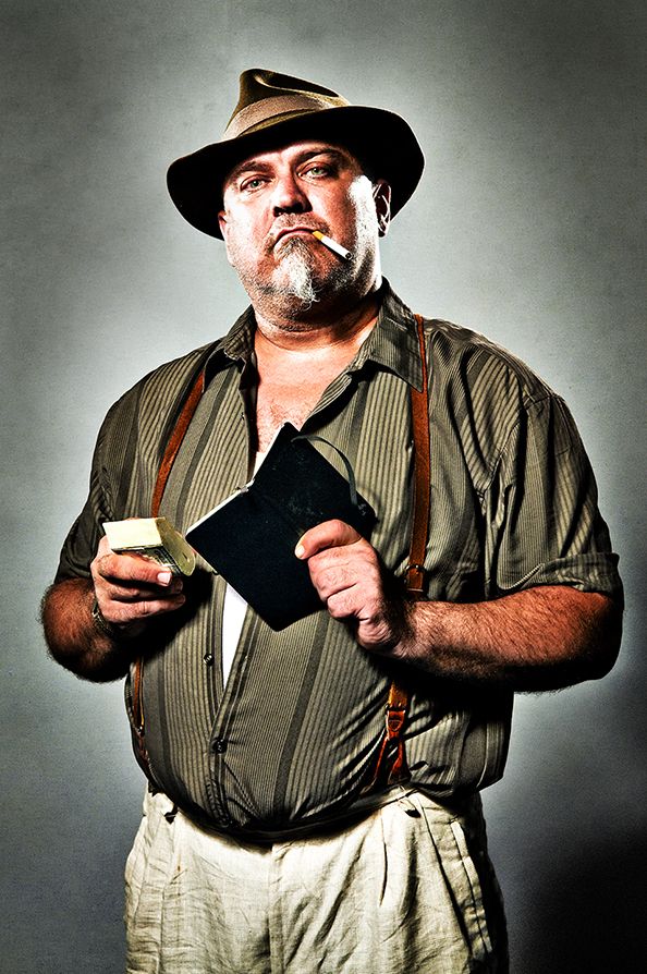 An older man in suspenders and hat poses confidently against a gray backdrop while holding a leather wallet.