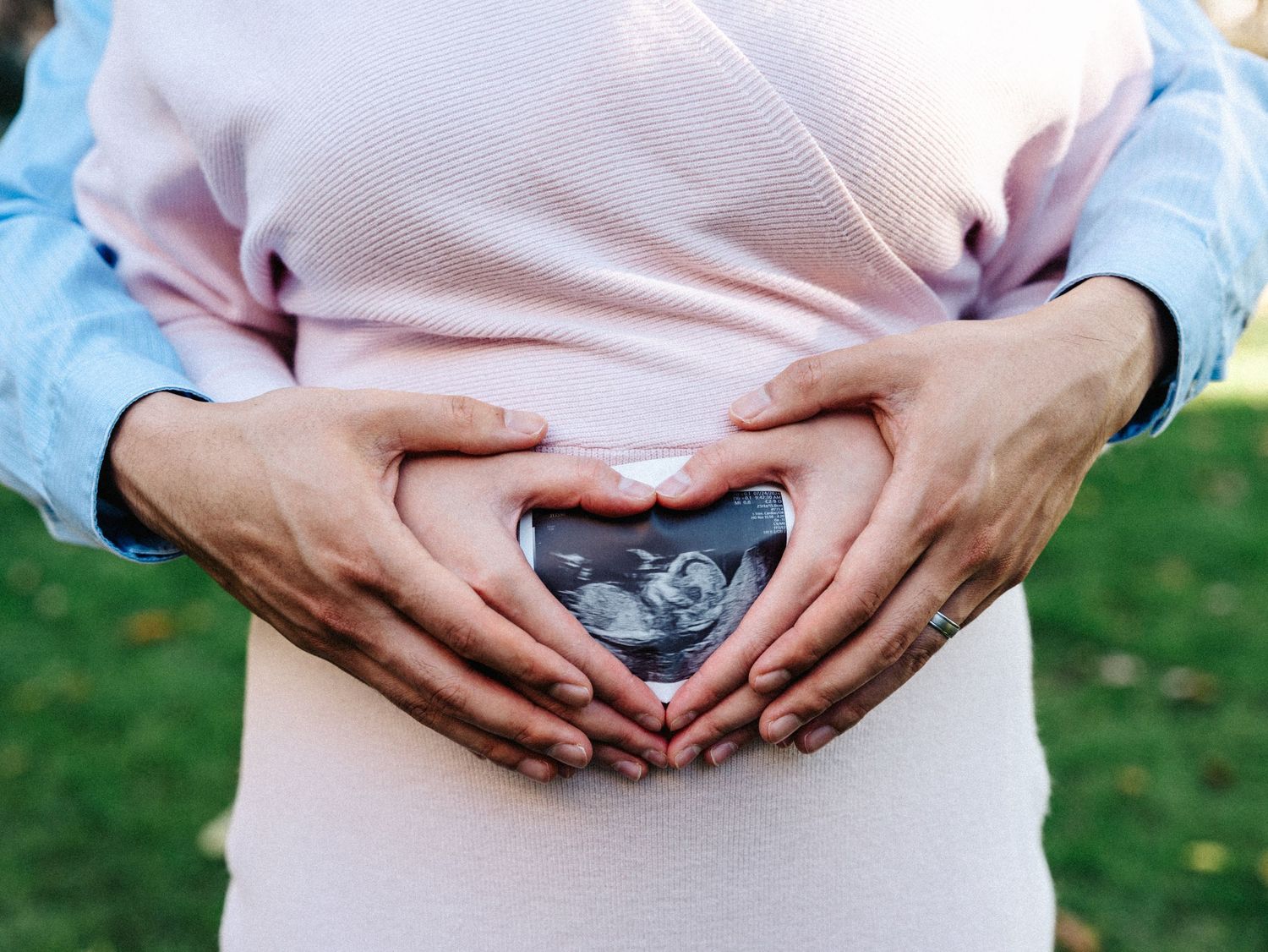 Hands shaped like a heart frame an ultrasound photo on a pregnant belly outdoors at Harvard campus.