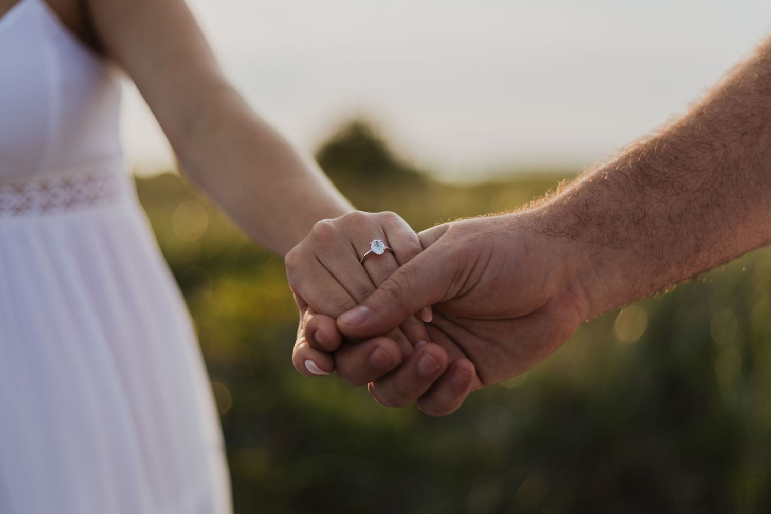 Two hands clasp tenderly together while wearing wedding rings, with one partner in a white dress in soft sunlight.