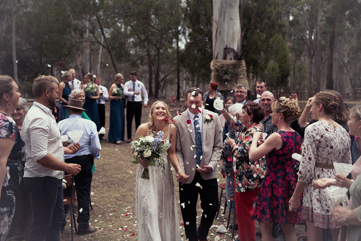 A newlywed couple is walking down an outdoor aisle surrounded by guests The bride is holding a bouquet and wearing a white dress, groom suit