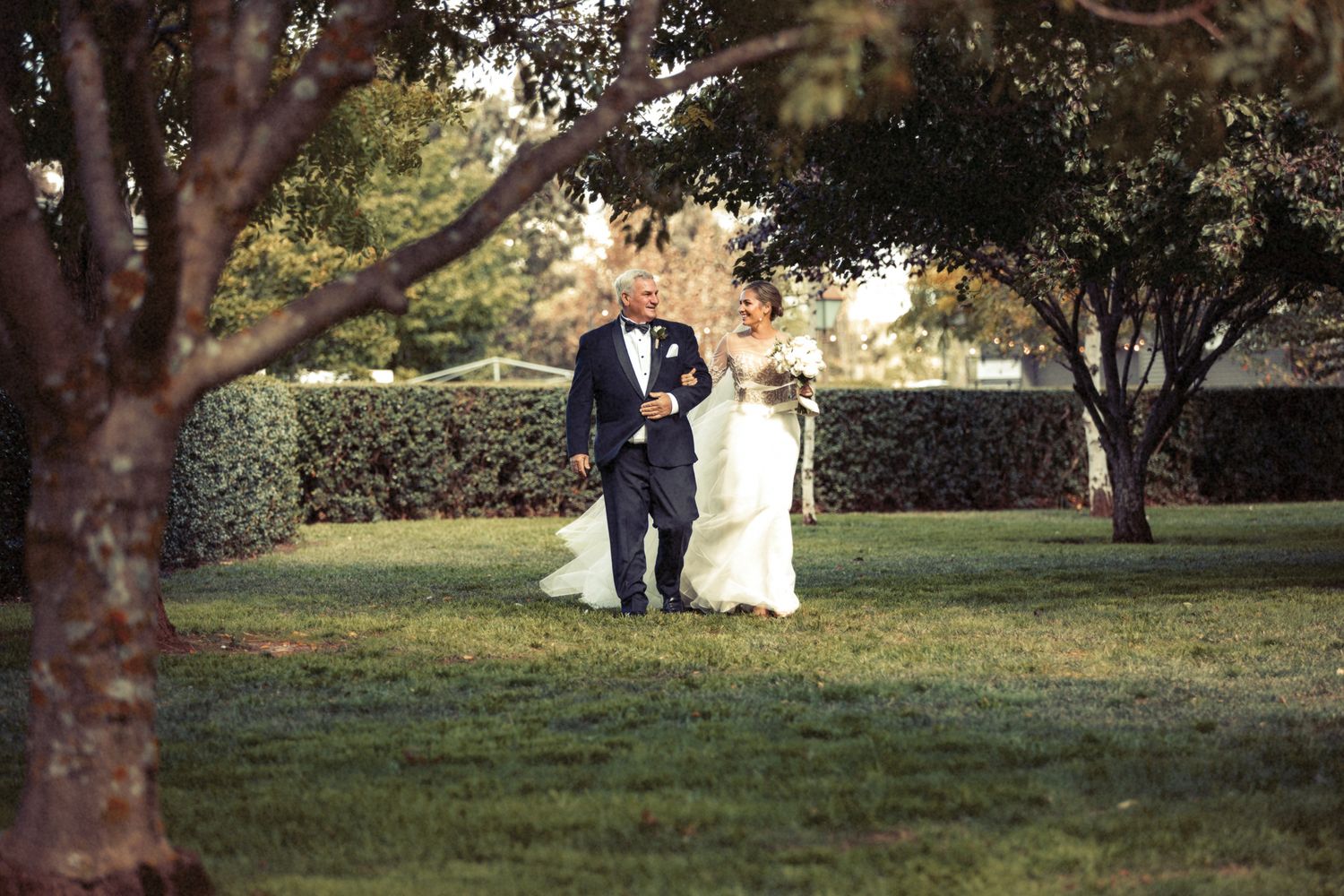 A bride and an older man are walking together on a grassy area surrounded by trees.