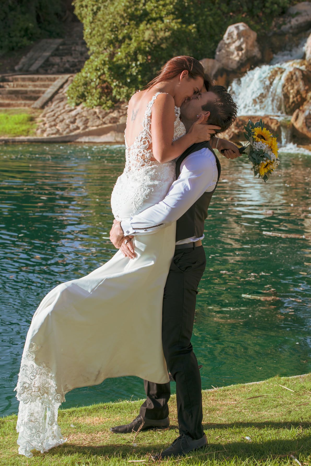 Romantic wedding pose by waterfall with bride holding sunflower bouquet.